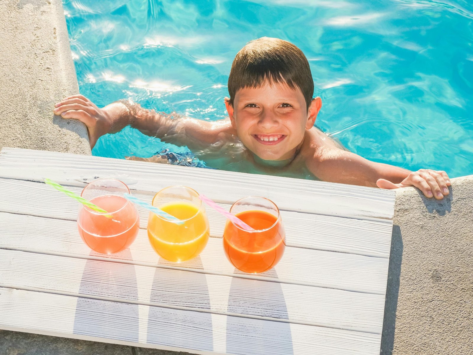 Smiling child in a swimming pool with three colorful drinks on a sunny day.